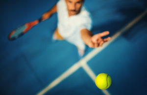 Take Advantage of Spring Conditions Closeup top view of a male tennis player hitting a ball during a serve. It's played on blue hard court surface just like at US open. Very shallow focus, only tip of the racket and the ball are in focus.
