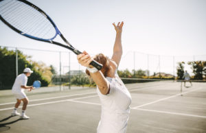 Daily Doubles Observations Rear view of a woman serving the ball while playing a mixed doubles tennis match. Men and women playing tennis outdoors on a sunny day.