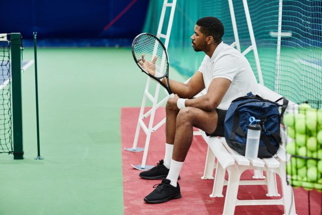 Black Tennis Player at Practice Side view portrait of professional tennis player holding racket while taking break on bench in court, copy space