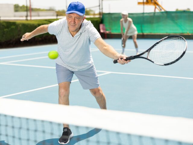 Portrait of concentrated elderly man playing tennis with friend on court.