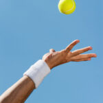 Serving ball. Close-up of male hand in wristband throwing tennis ball against blue sky