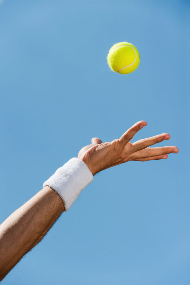 Serving ball. Close-up of male hand in wristband throwing tennis ball against blue sky Serving ball. Close-up of male hand in wristband throwing tennis ball against blue sky