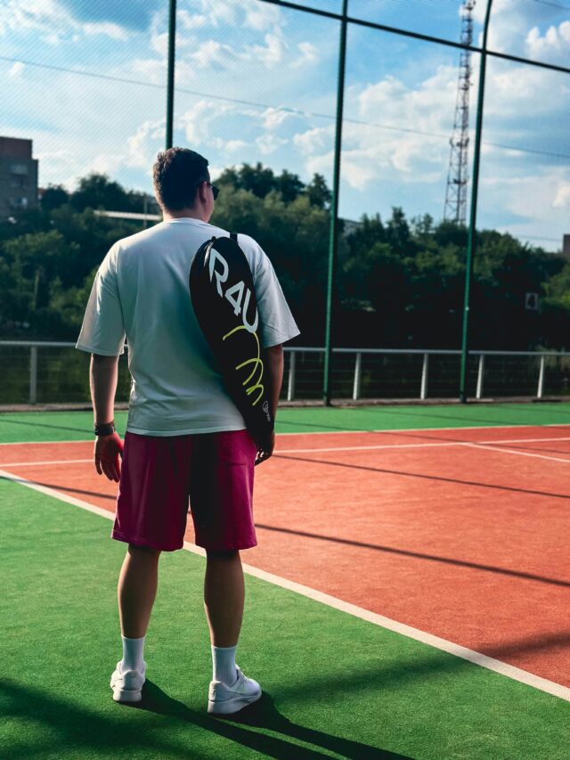 denis-zelenykh-U342klzF4TI-unsplash Male tennis player standing on a court with a racquet bag over his shoulder.